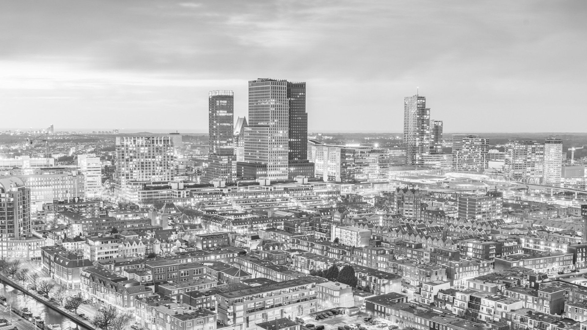 Een zwart-wit luchtfoto van een uitgestrekt stedelijk landschap met wolkenkrabbers die boven dichtbebouwde woonwijken uittorenen.