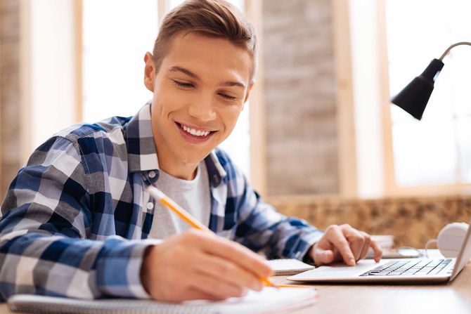 Handsome smiling fair-haired boy working on his laptop and writing