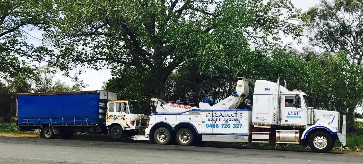 A White Tow Truck Towing a Blue Truck Under a Large Tree — Haigh Truck & 4WD Repairs In Orange, NSW