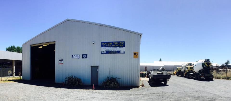 A white building with an open garage door, vehicles are parked outside on a gravel lot. Blue sky in the background.