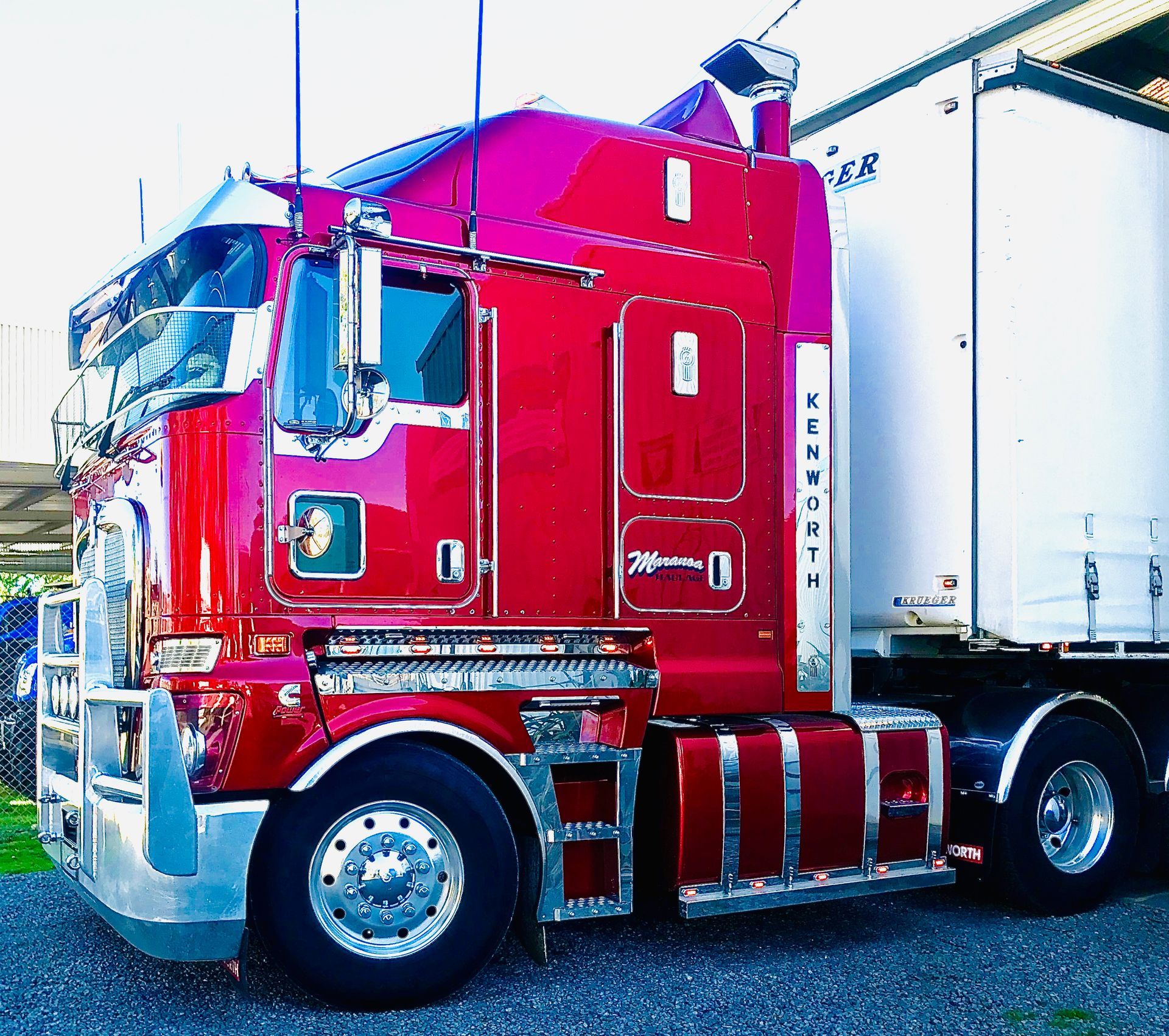 Red semi-truck with chrome details, parked on gravel. Trailer attached, white with blue lettering.