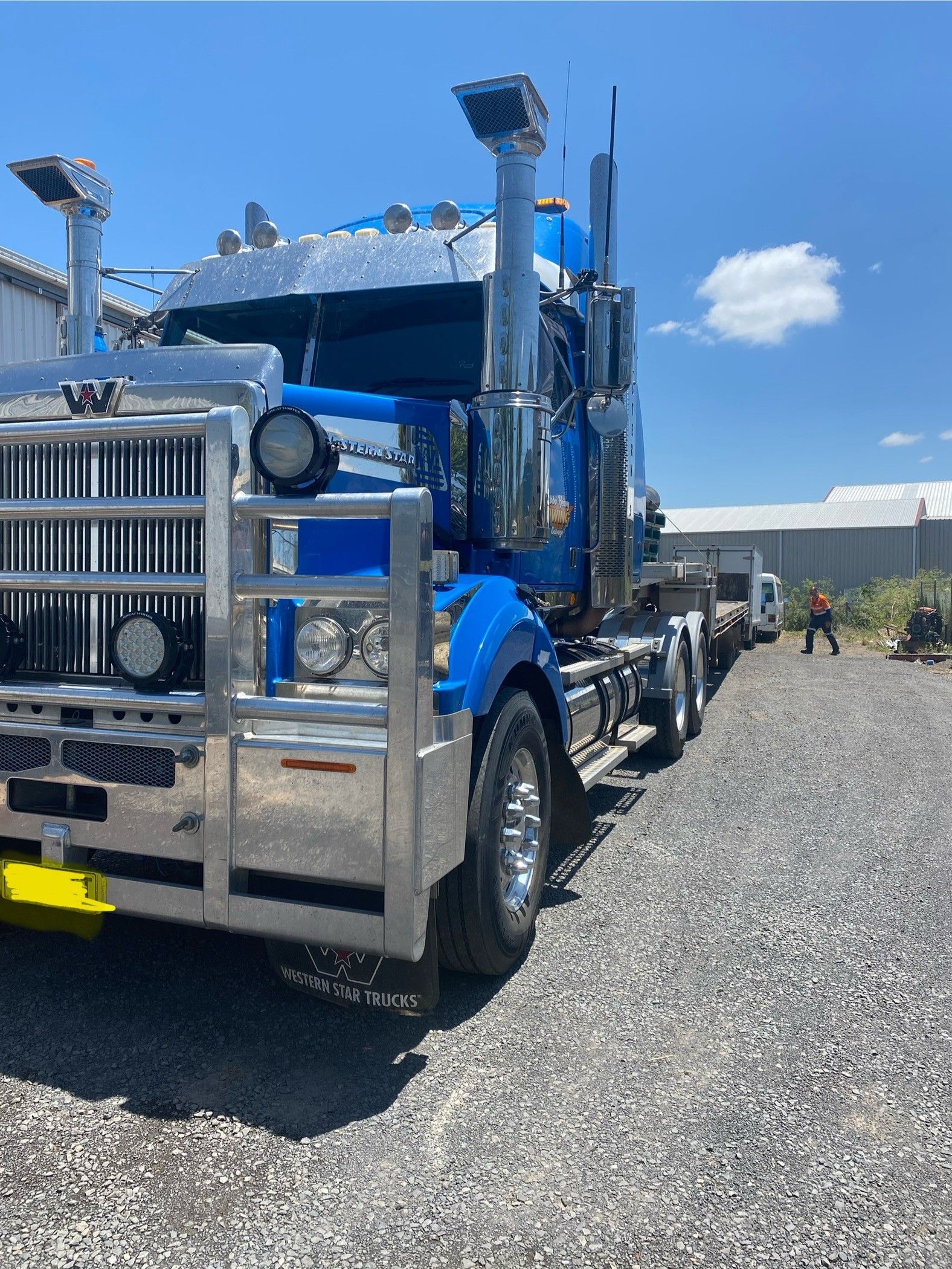 Blue semi-truck with chrome details parked on gravel under a clear sky. A person stands in the distance.