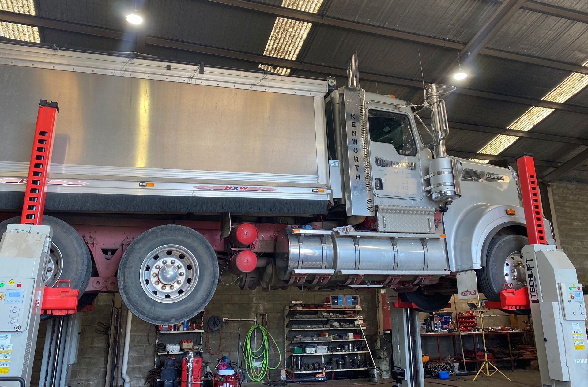 Semi-truck lifted in a garage for repairs. Red lifts support the truck. Silver truck and red accents.