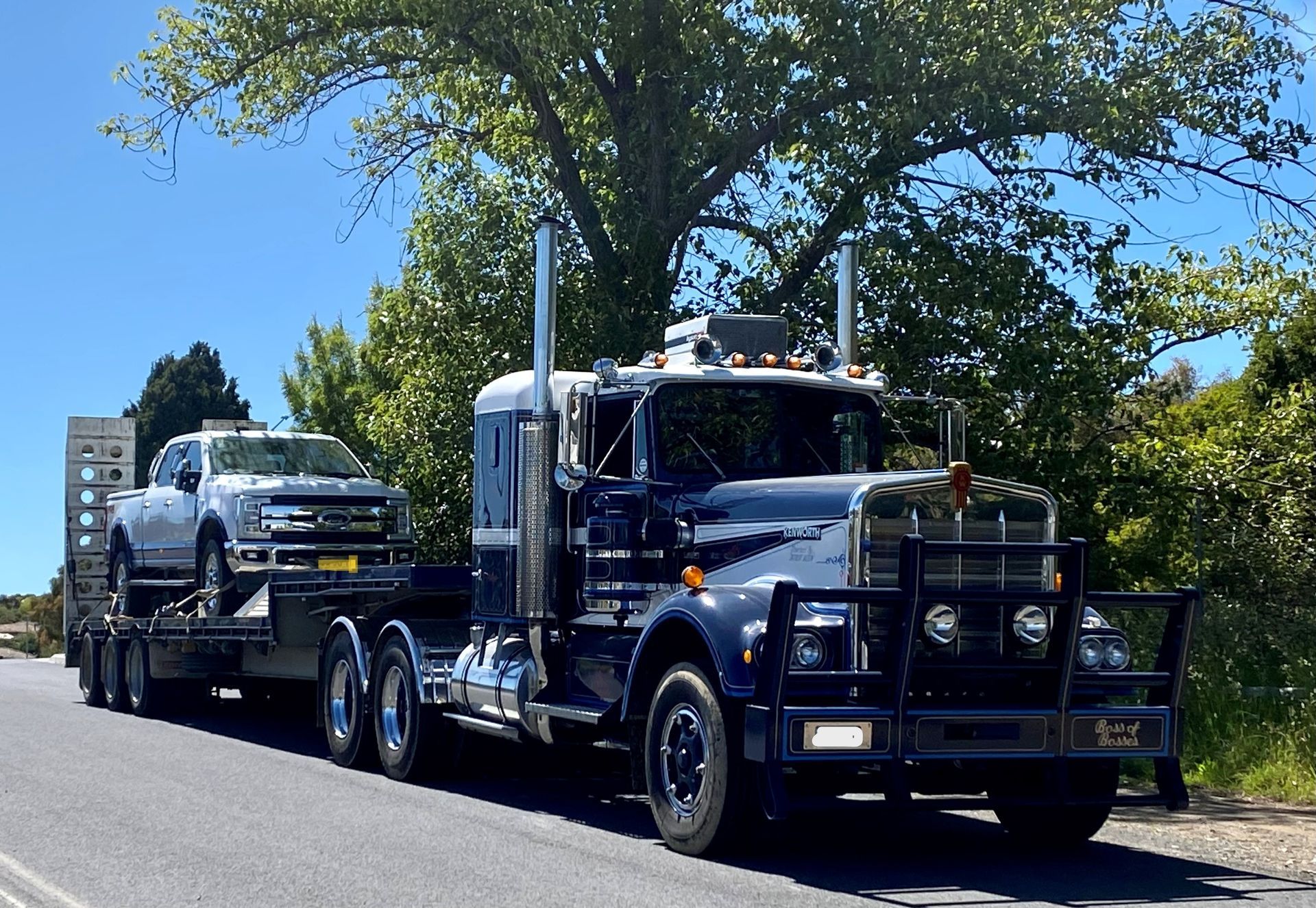 Dark blue semi-truck towing a flatbed trailer carrying a white pickup truck on a sunny road.