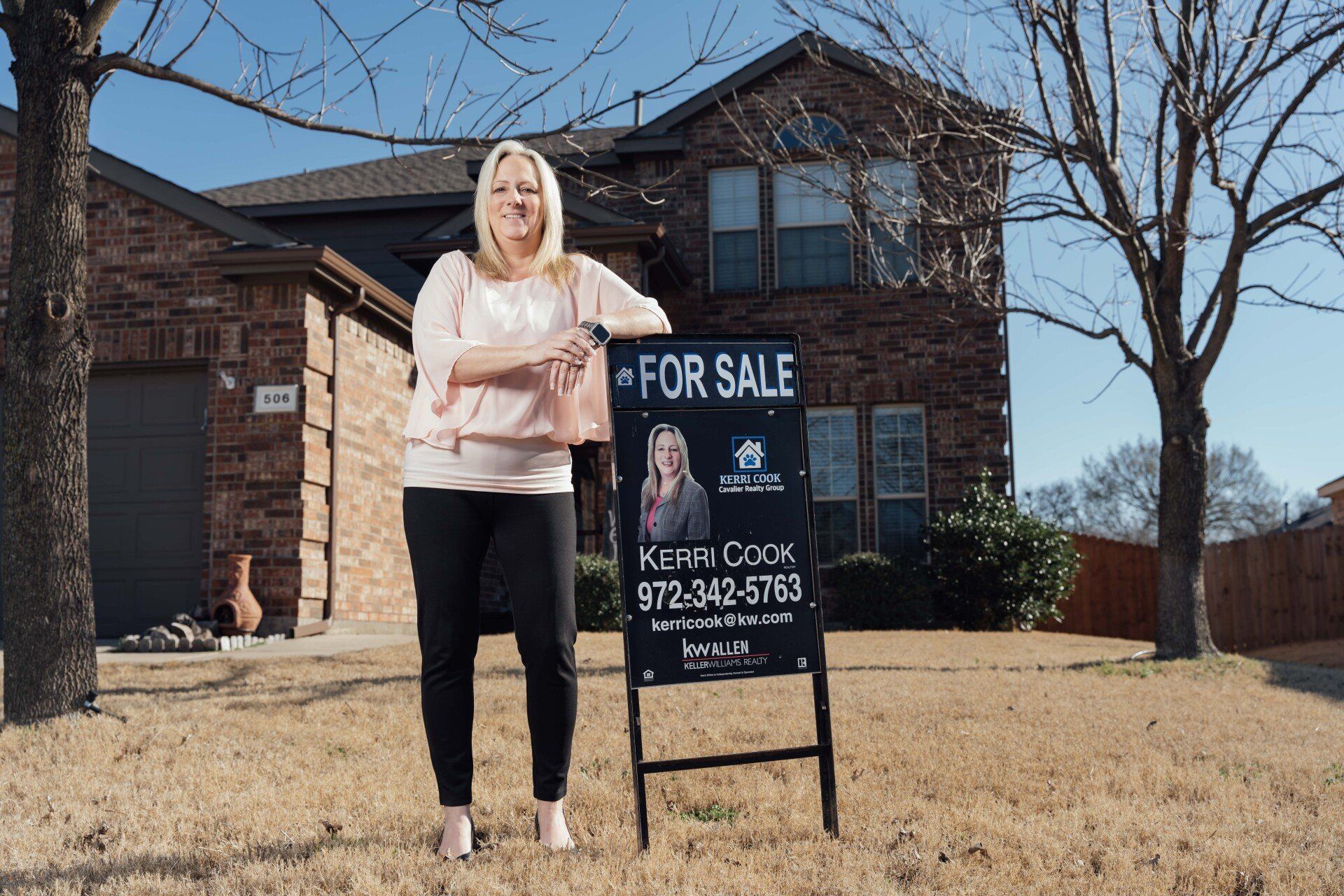 A woman is standing in front of a house holding a for sale sign.