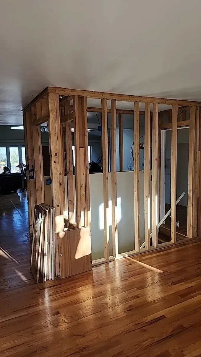 Interior view of partially demolished wall with exposed wooden frame in a room with wood flooring.