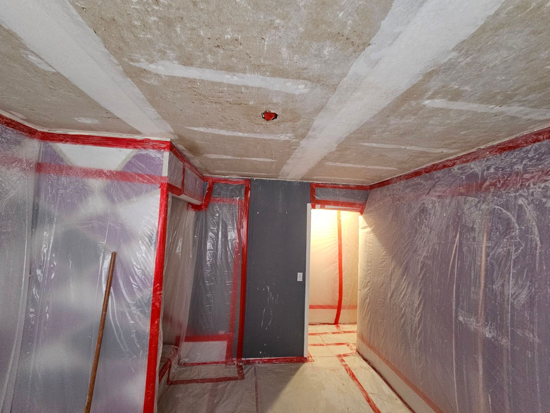 Room sealed with plastic sheeting, red tape, ready for renovation. Gray door and ceiling visible.