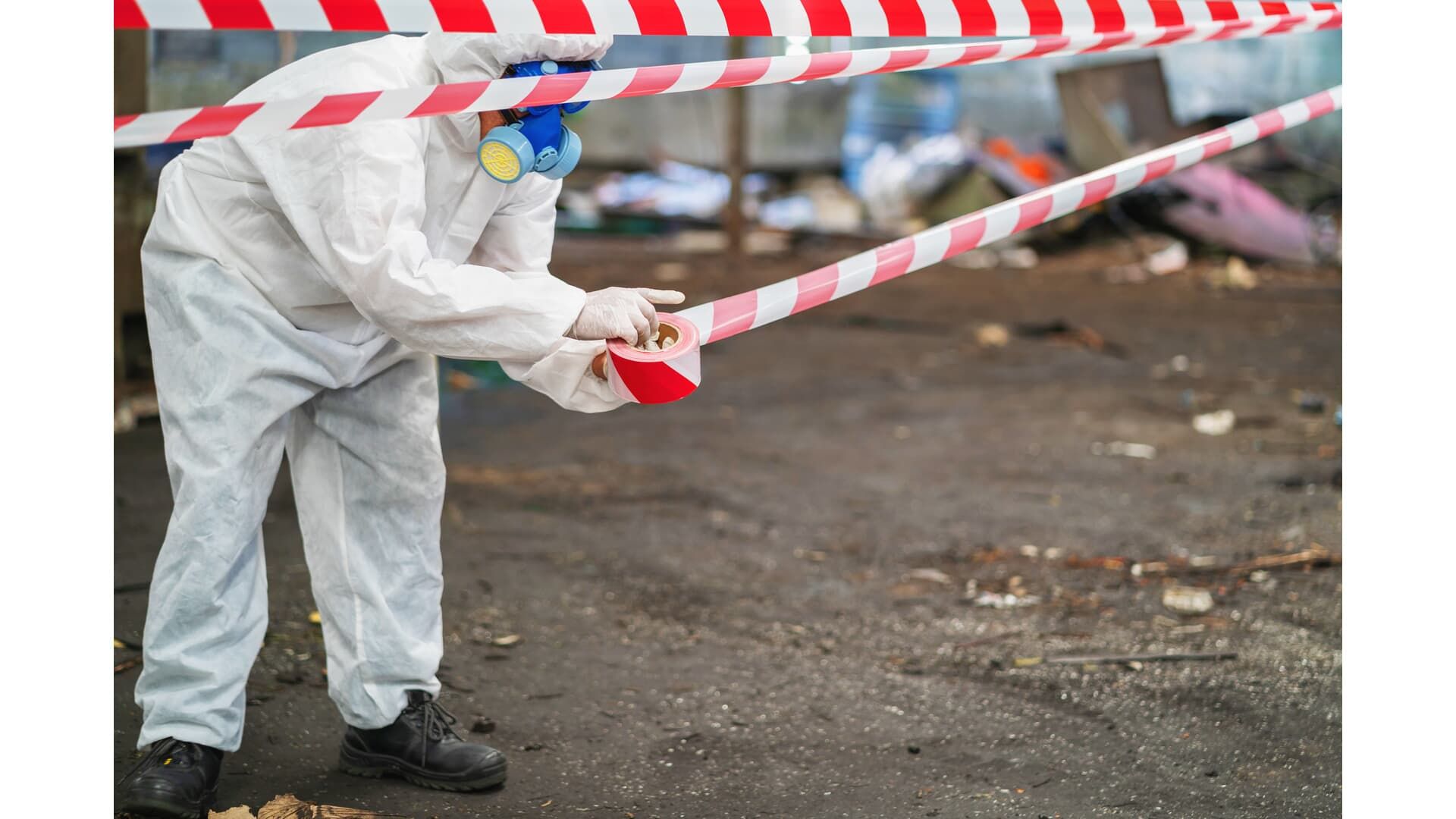 Person in hazmat suit securing red and white barrier tape in a debris-filled area.
