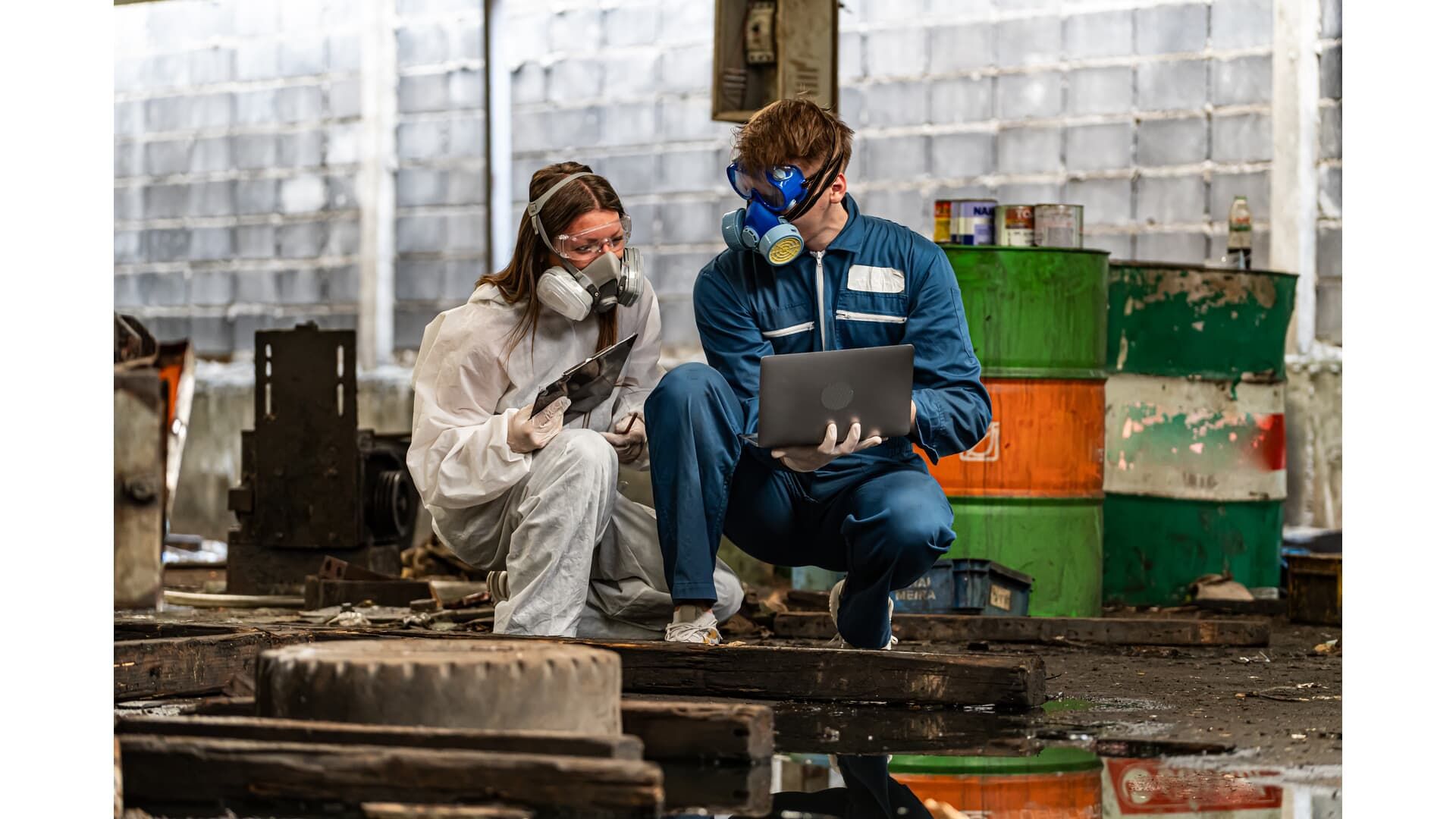 Two people in protective gear examining a laptop in a hazardous industrial setting.