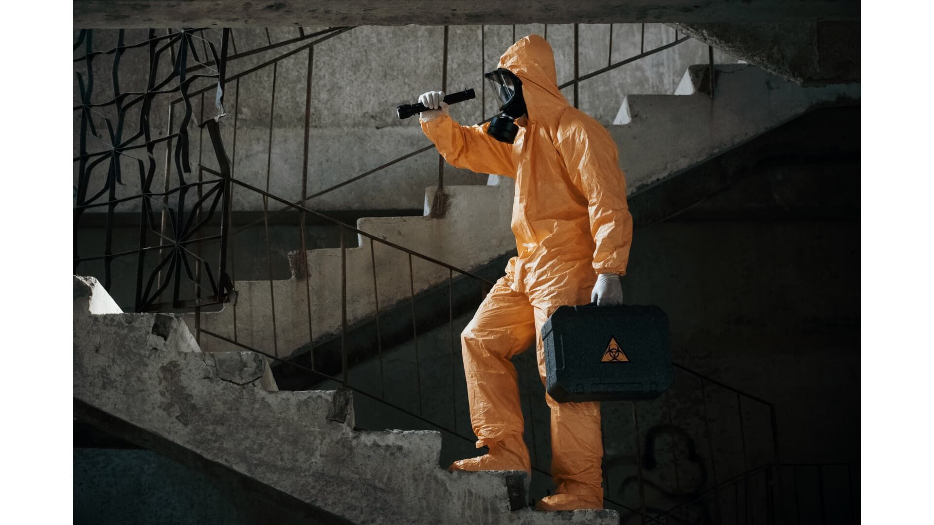 Person in hazmat suit with a briefcase and flashlight climbing stairs in a dimly lit, concrete building.