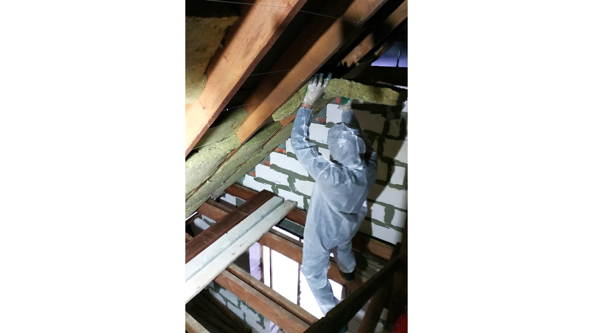 Person in protective suit installing insulation in an attic with exposed beams and brick wall.