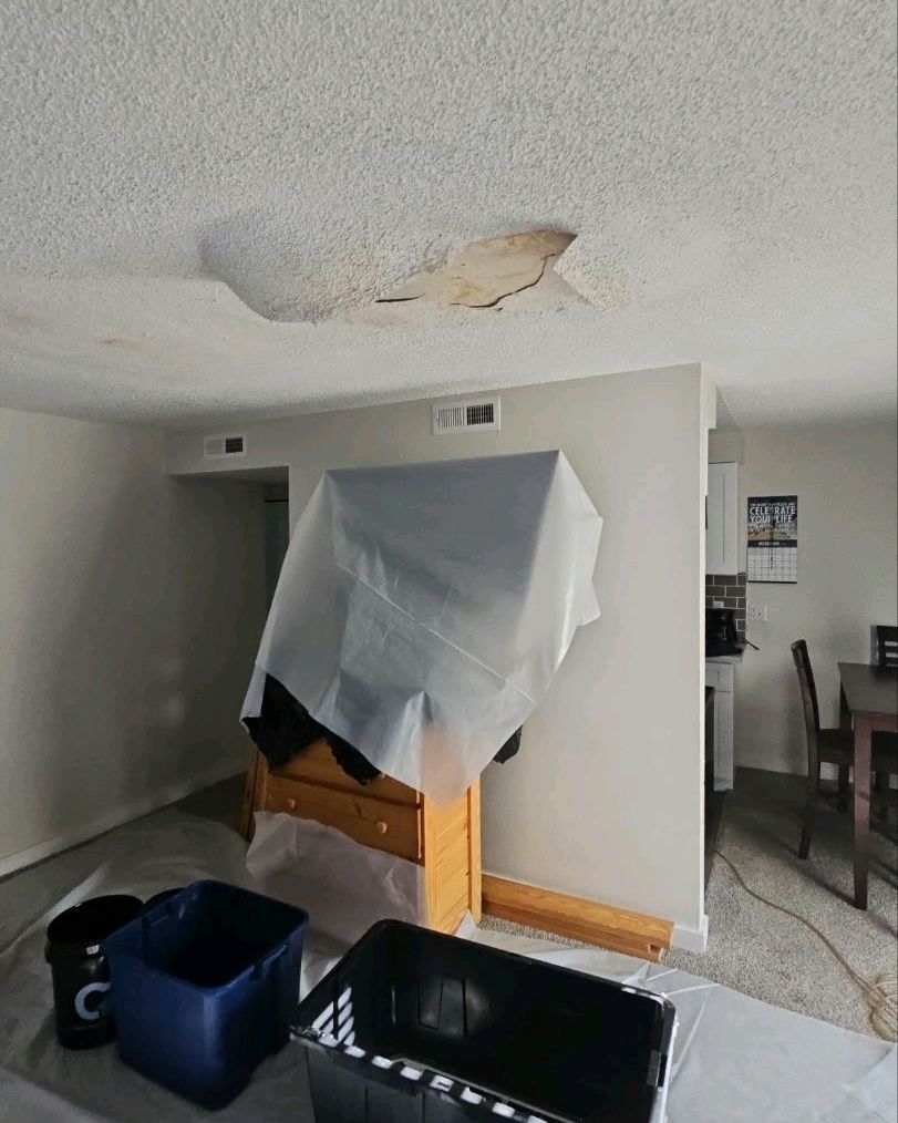 Ceiling damage in a room with furniture covered in plastic, and buckets on the floor.