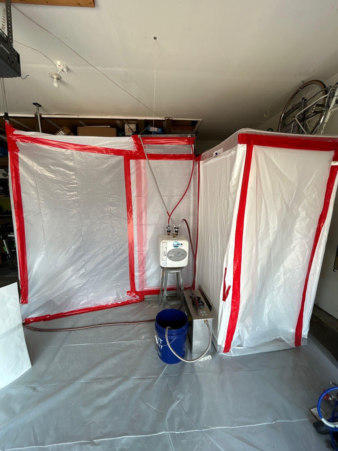 Spray booth in a garage, made with red-trimmed plastic sheeting. Equipment in the center, a blue bucket.