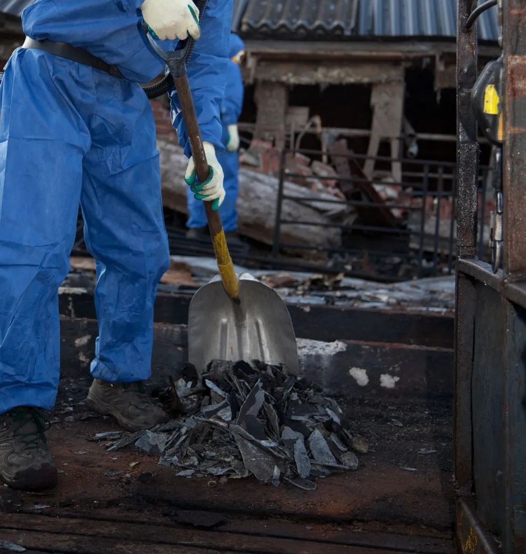 Person in blue coveralls shoveling debris from a charred structure.