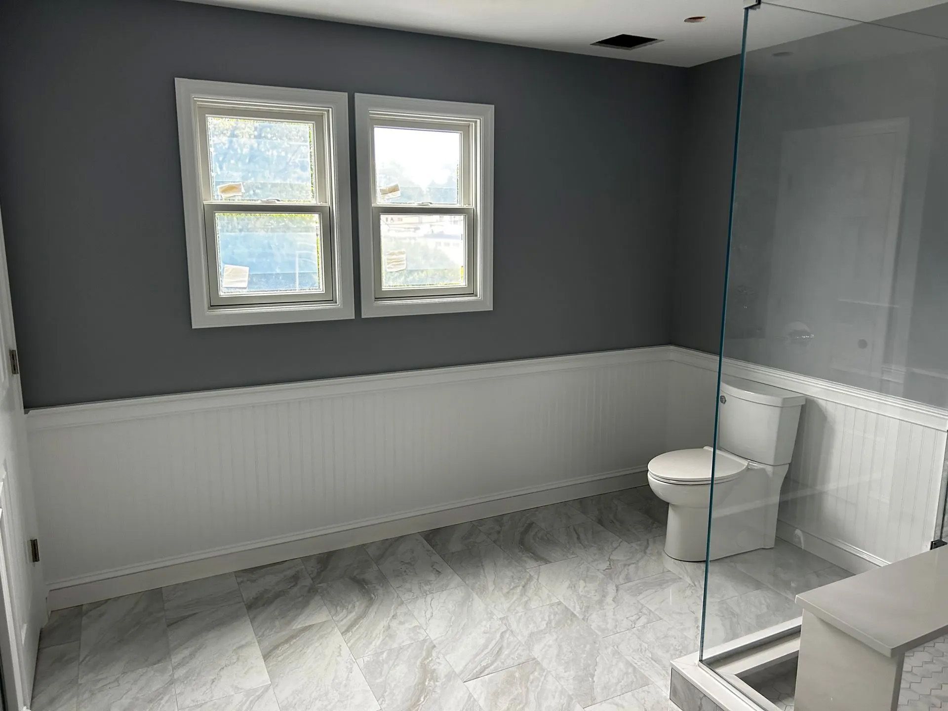 A bathroom with a toilet , shower and two windows.