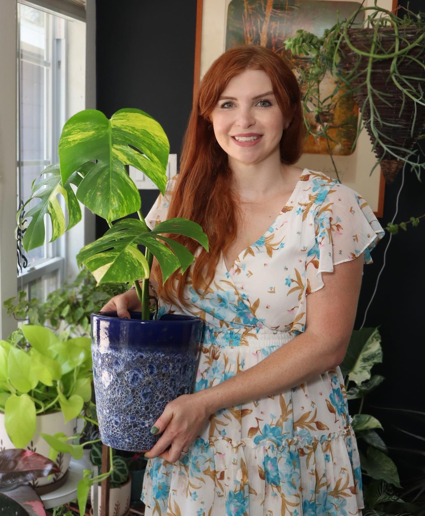 Woman holding a potted Monstera plant, smiling, in front of a window and other plants.