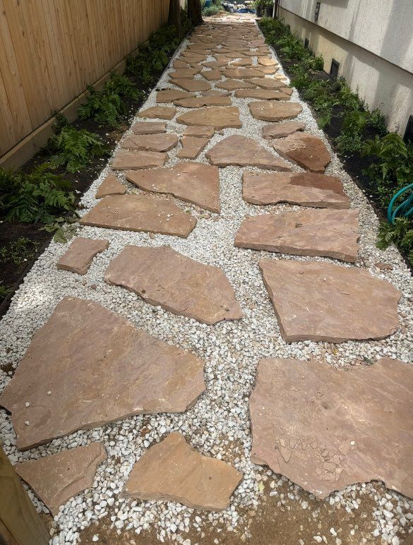 Flagstone walkway through a narrow outdoor space, bordered by plants and gravel.