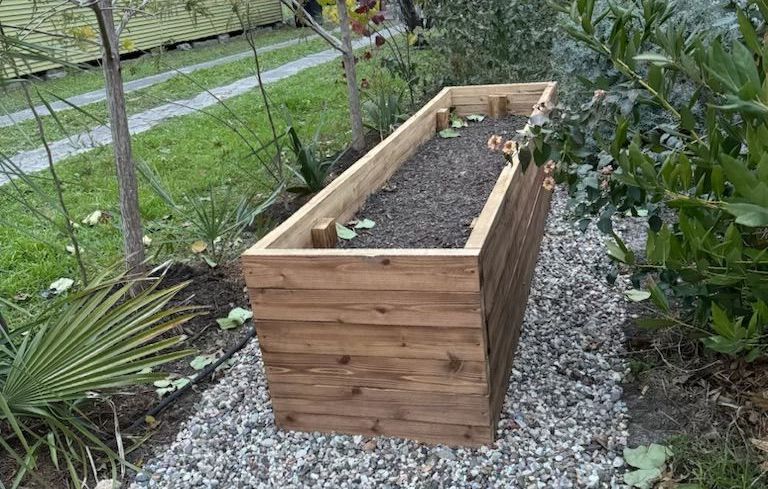 Wooden rectangular raised garden bed filled with soil; surrounded by gravel, plants, and grass.