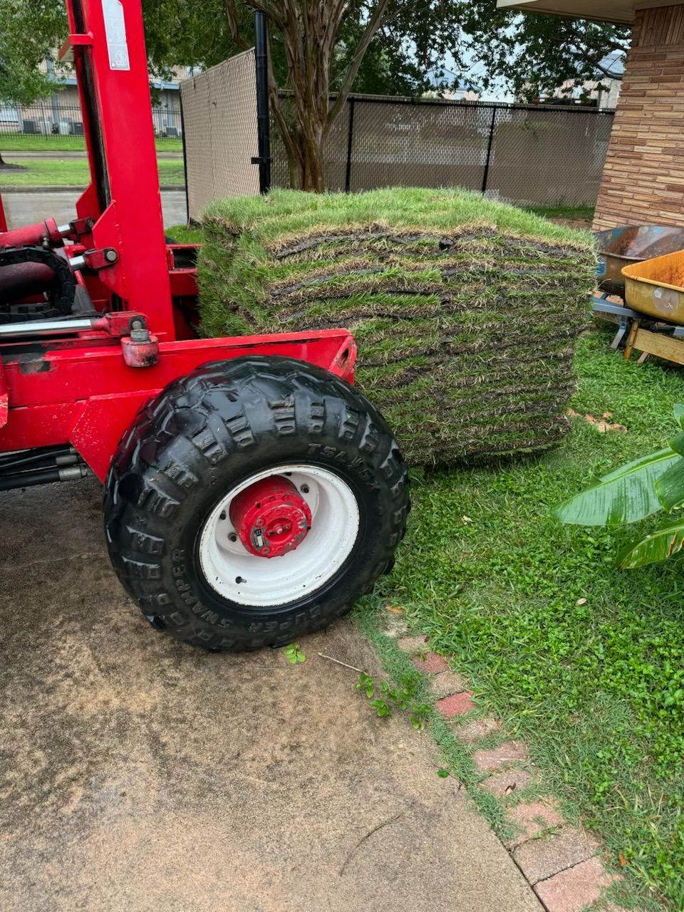 Red forklift holding a stack of sod on green grass next to a brick wall.