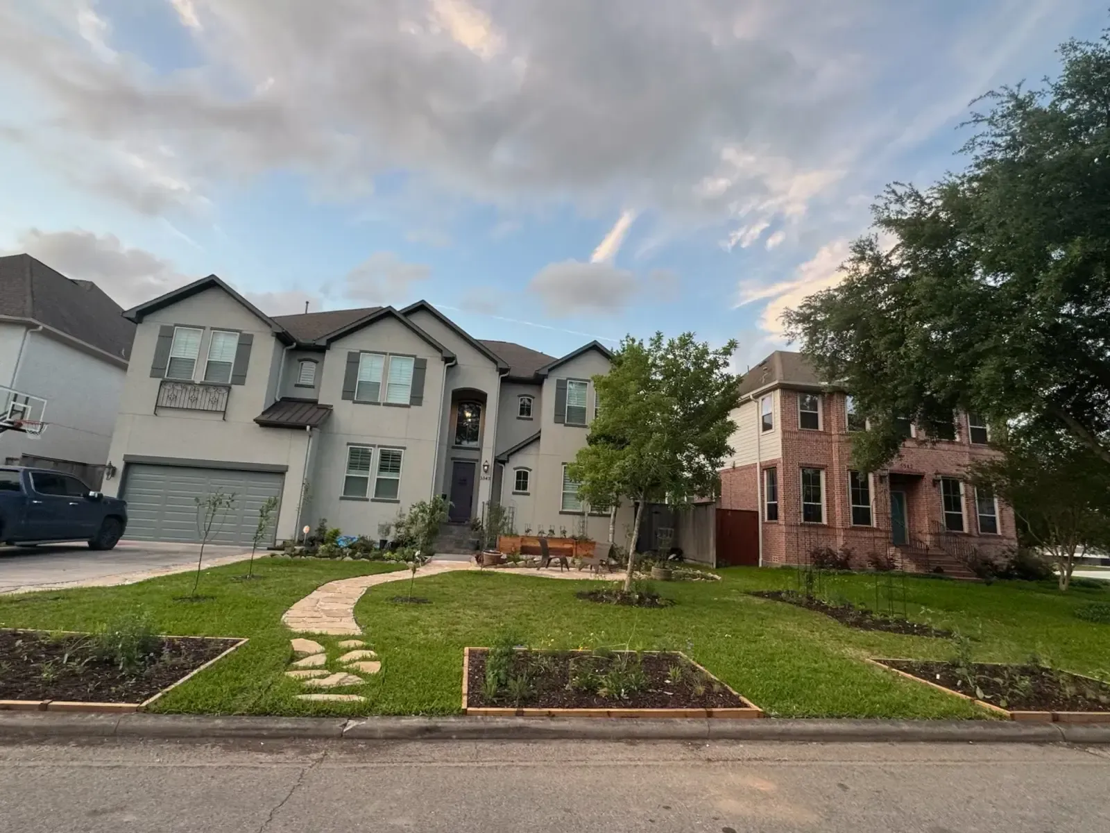 Two-story house with light-colored stucco, gray roof, and a lawn with flower beds. Cloudy sky.