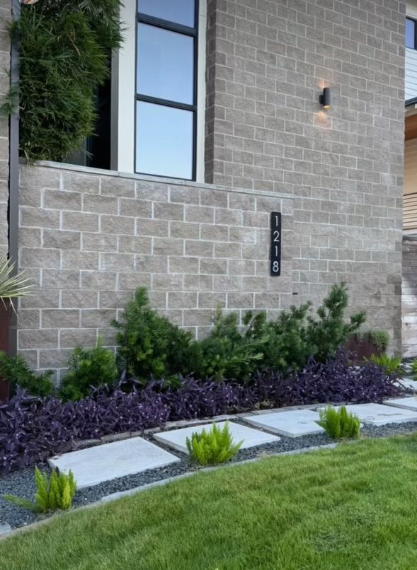 Gray brick building facade with address 728, greenery, purple plants, and a stone walkway.