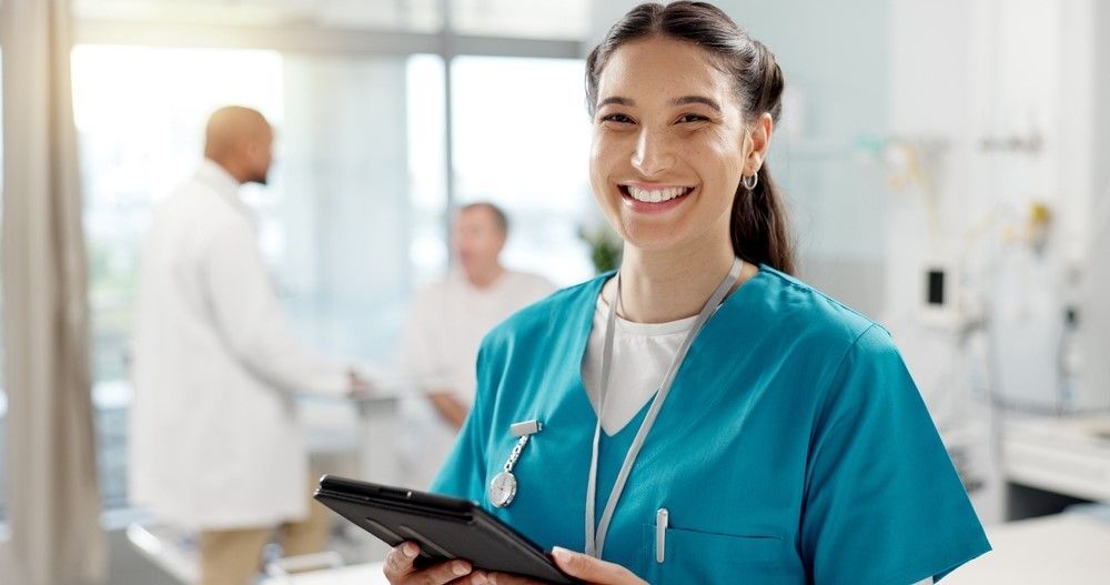 A nurse is smiling while holding a tablet in a hospital room.