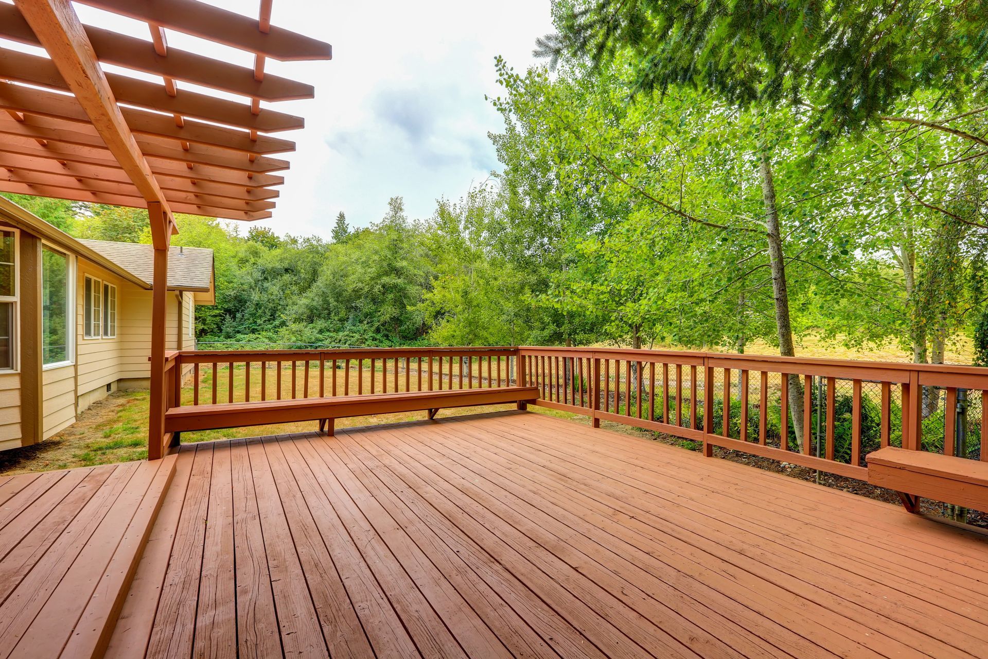 An empty wooden deck with a pergola and trees in the background.