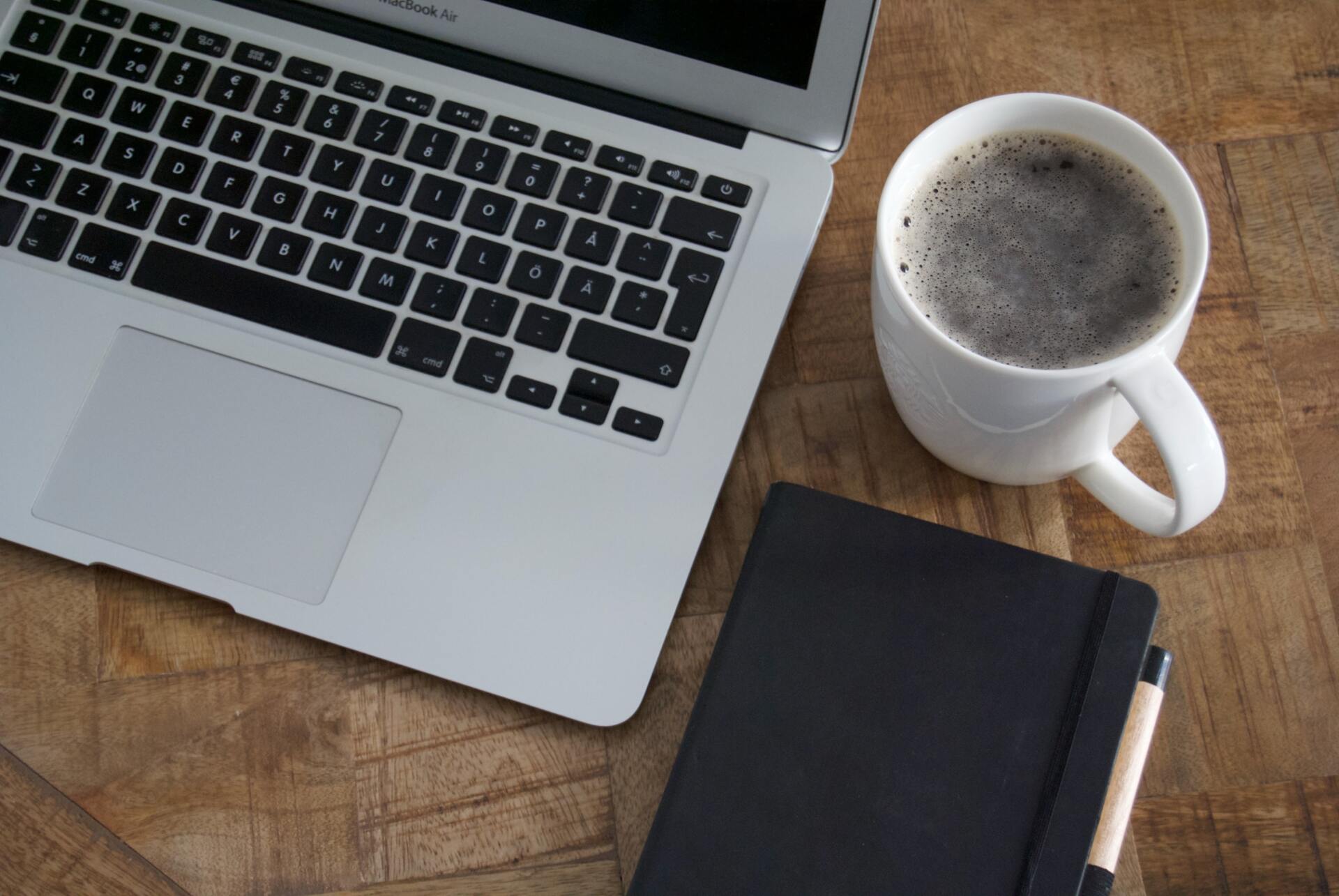 An image with an open laptop, close black notebook with pen, and a cup of black coffee in a white mu