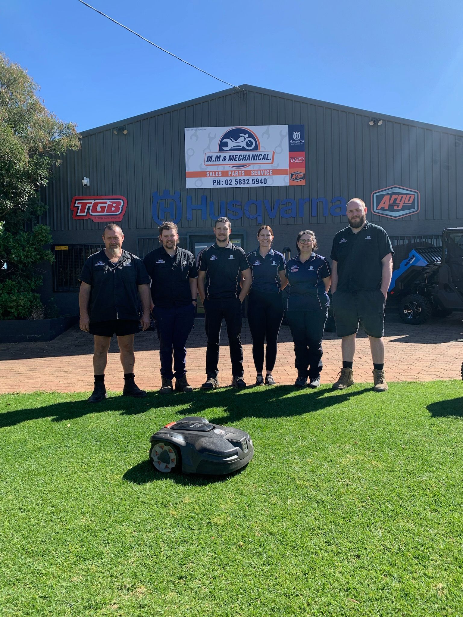 Group of people standing in front of a building with a lawnmower, wearing black shirts on a sunny day— M.M & Mechanical in Dubbo, NSW