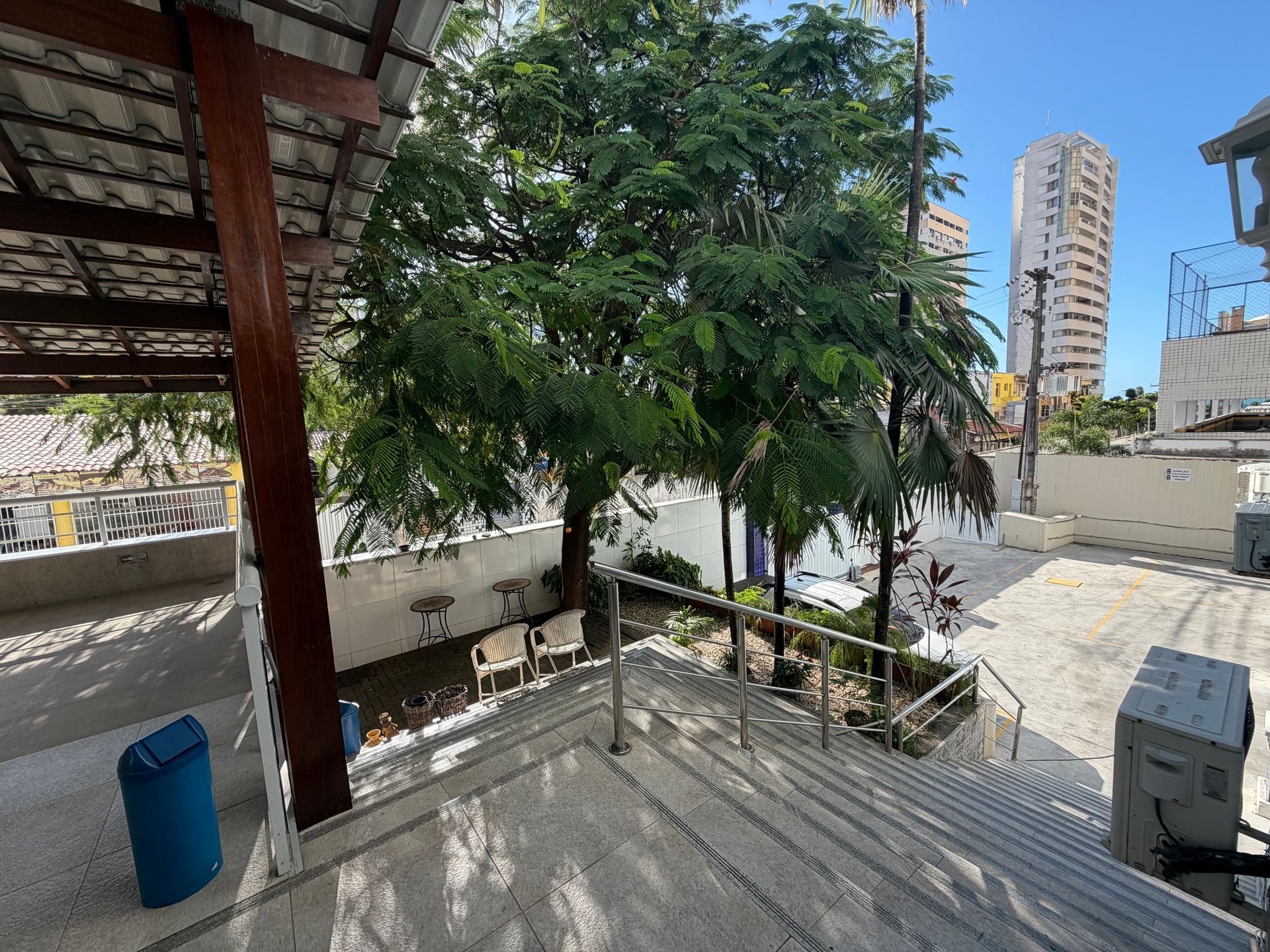 Terraço na cobertura com escadas, uma árvore frondosa, um barril azul e prédios da cidade ao fundo.