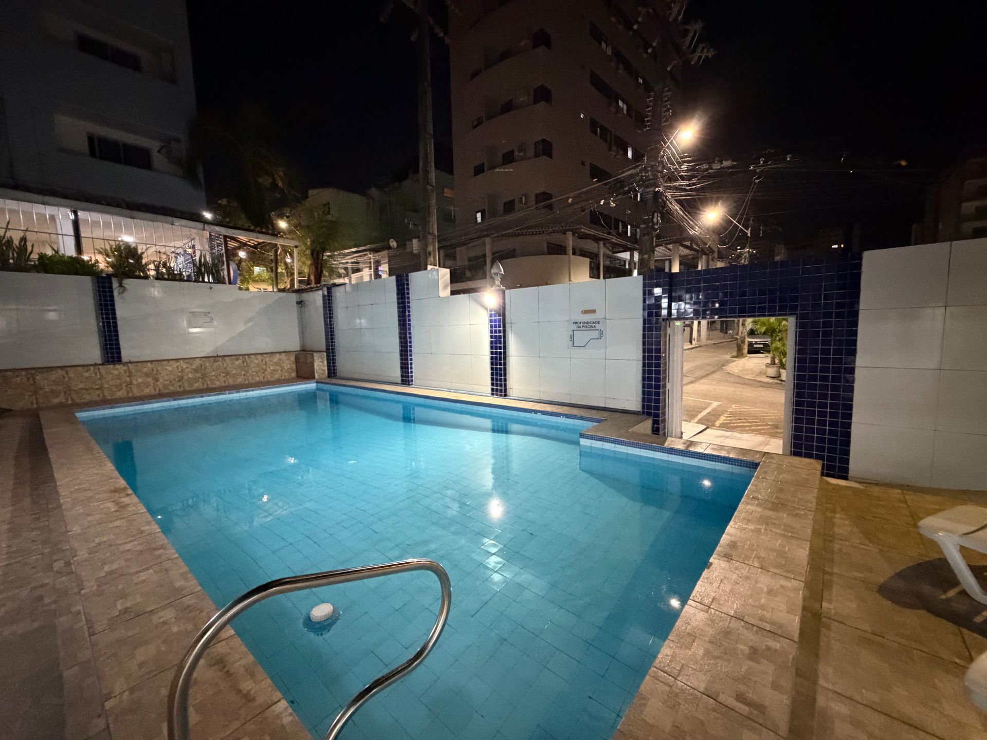 Piscina iluminada no quintal à noite, com água azul, deck de azulejos e prédios de apartamentos ao fundo.
