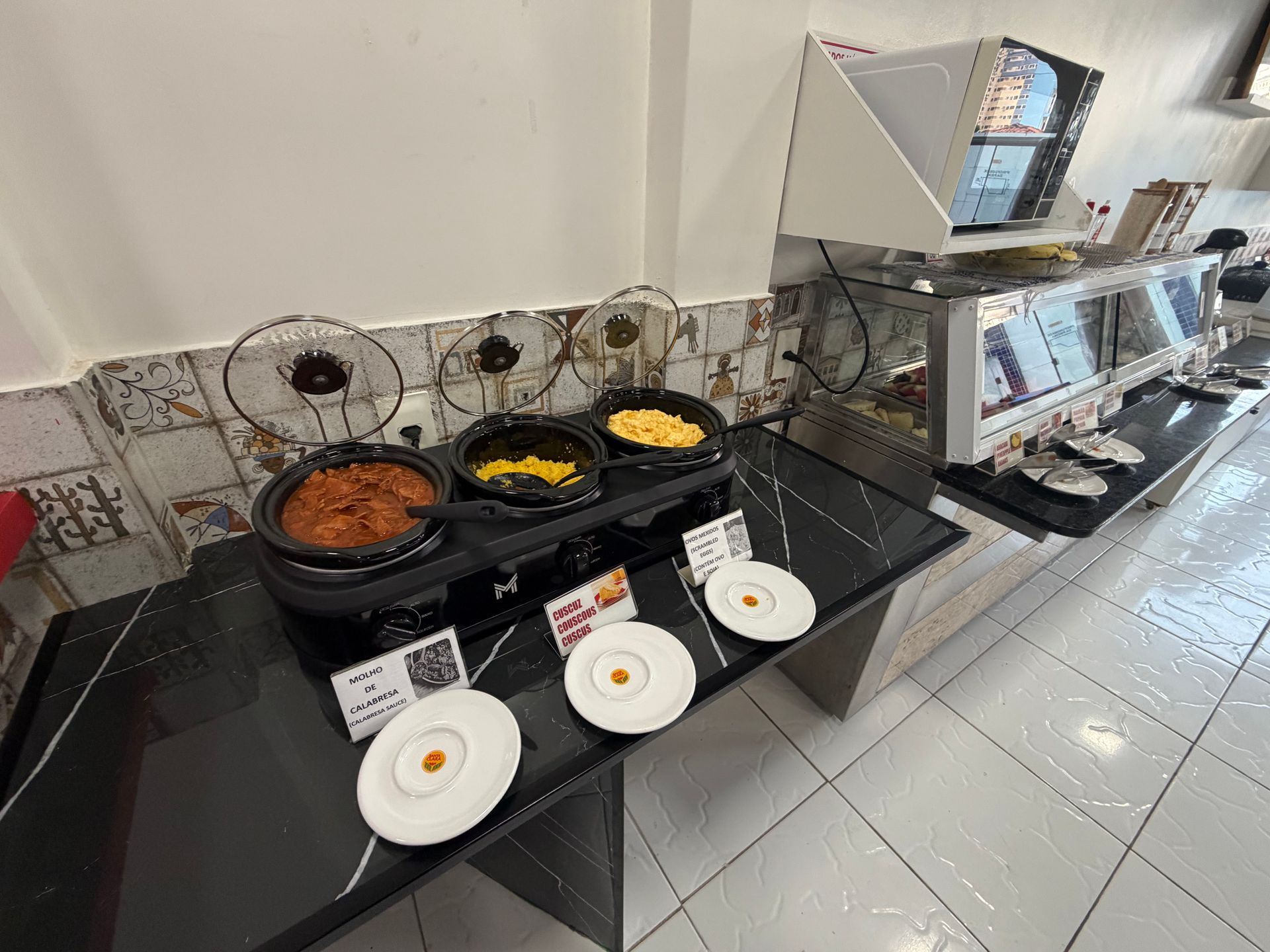 Balcão de buffet com pratos e travessas cobertos, e aquecedor de alimentos em aço inoxidável em uma área de refeições com piso de azulejo.