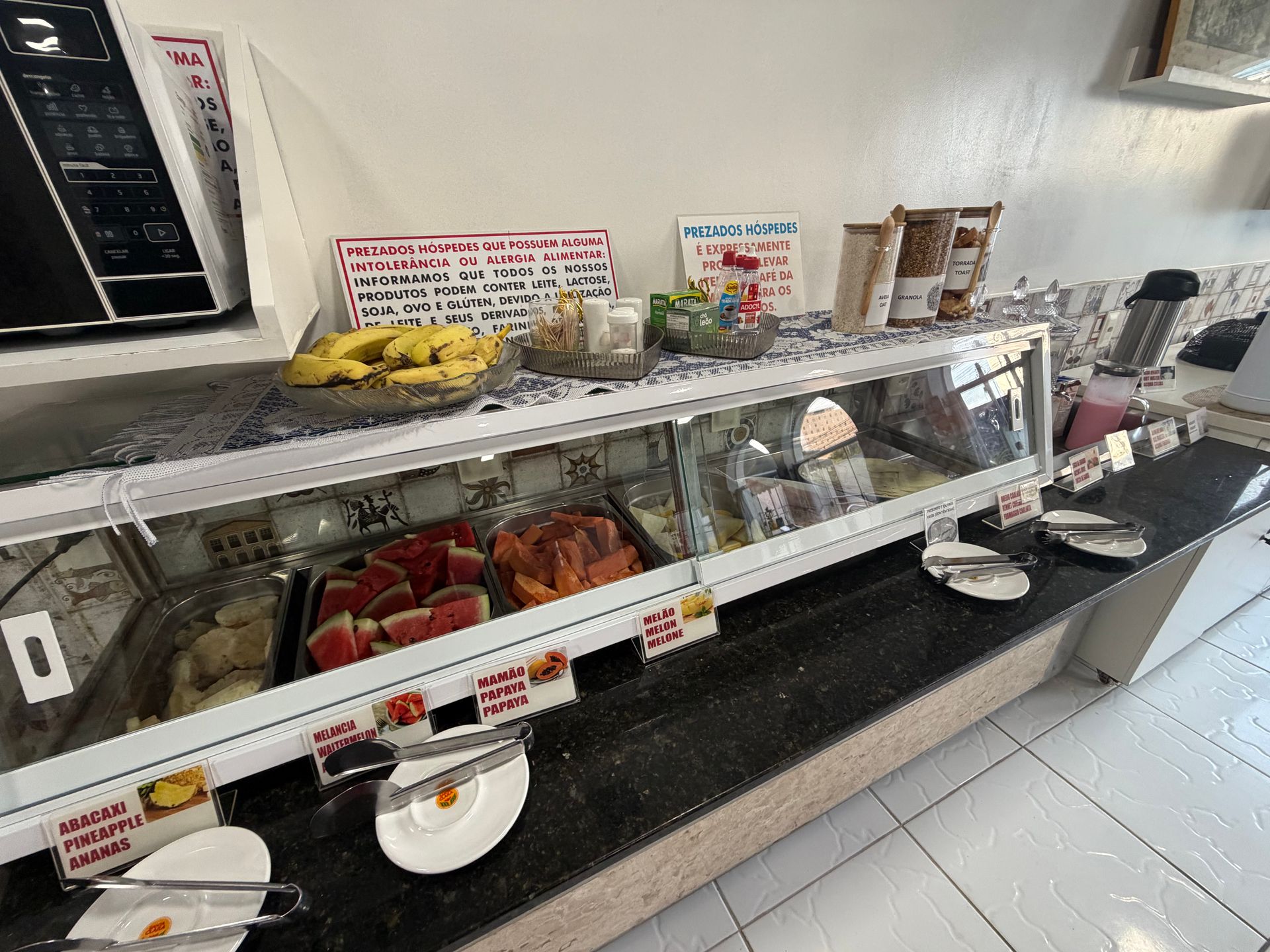 Buffet self-service com frutas, doces e pratos, protegido por uma divisória de vidro em uma área de refeições com piso de azulejo.