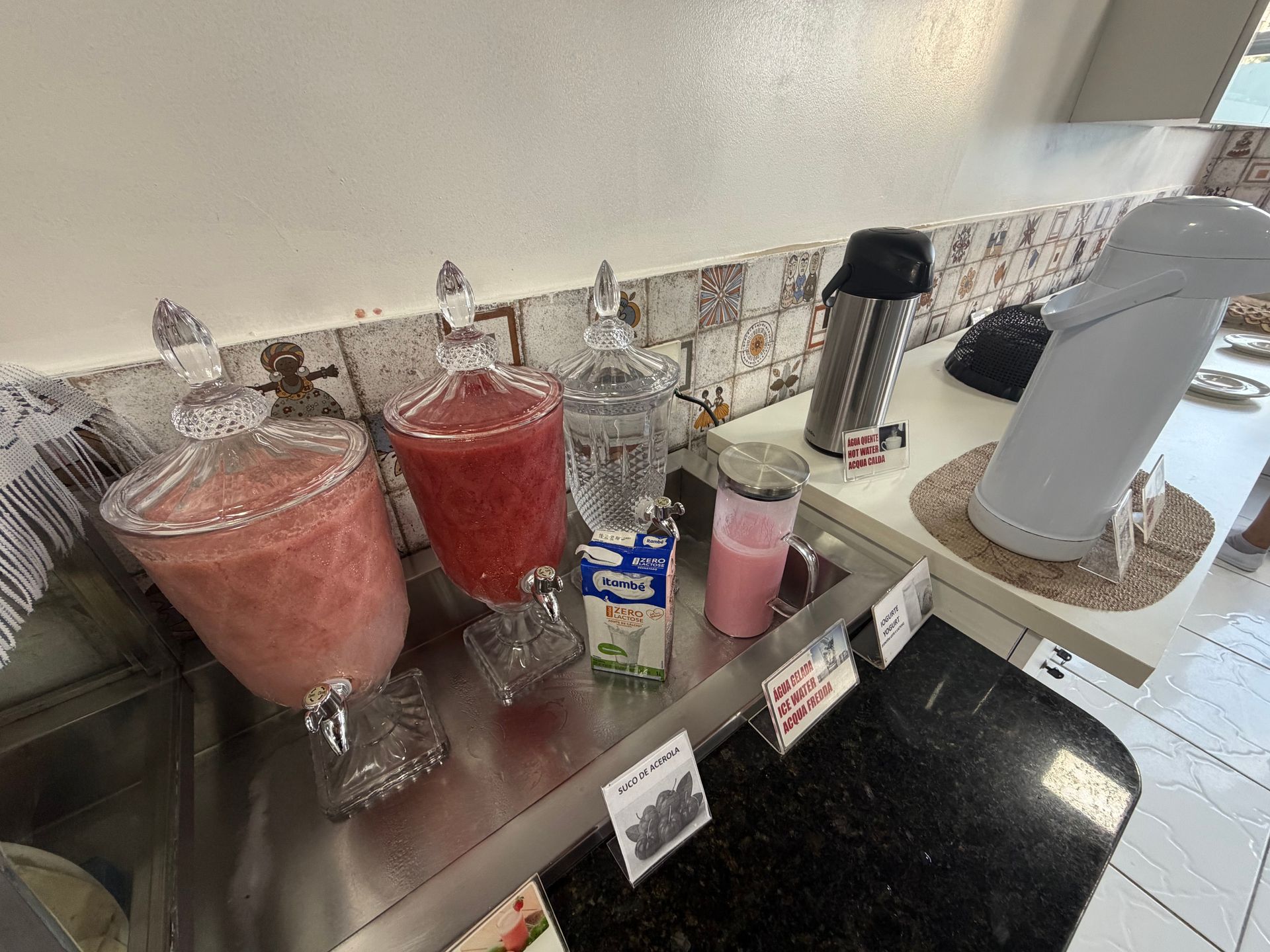 Estação de bebidas self-service com smoothies rosa, uma bebida vermelha, café preto e etiquetas em um balcão.