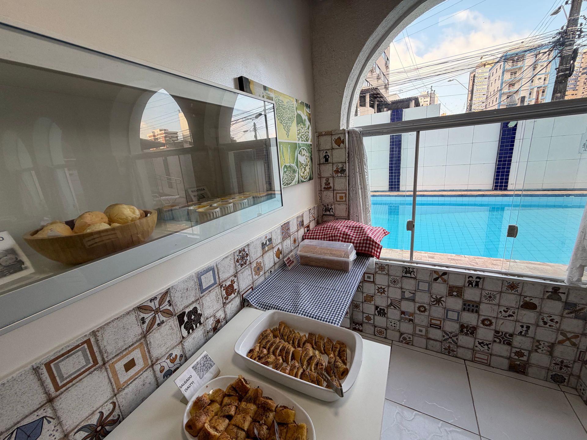 Área de bufê revestida com azulejos, com doces e pães ao lado de uma piscina azul vibrante, com vista através de uma janela em arco.