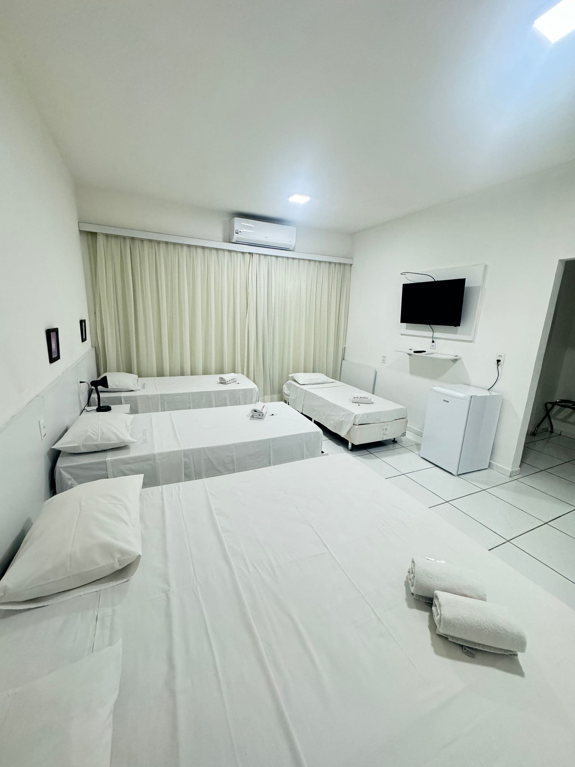 Quarto de hotel branco e luminoso com duas camas, cortinas, TV, cômoda e toalhas no chão.