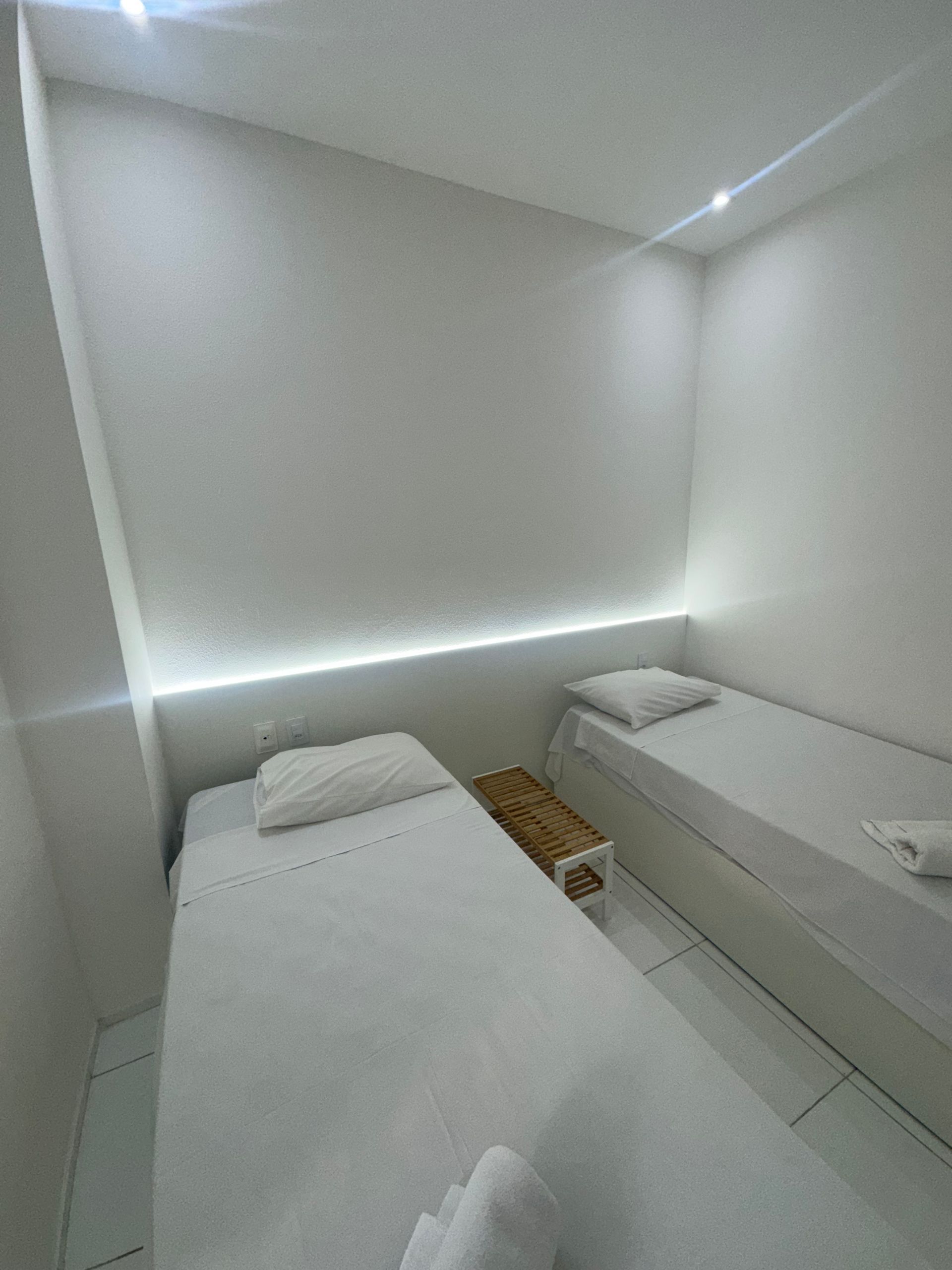Quarto pequeno e branco com duas camas de solteiro e um banco junto à janela.