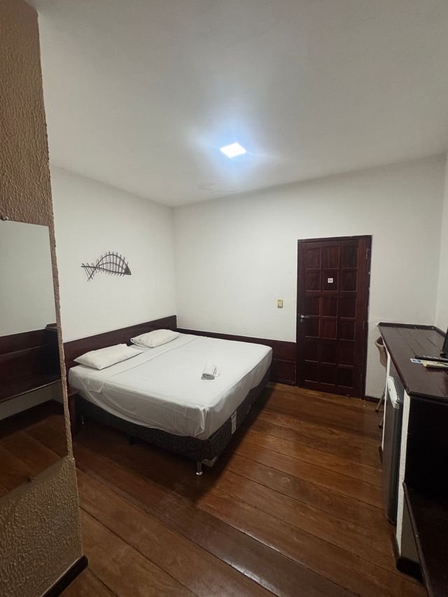 Quarto pequeno com cama, piso de madeira, porta escura e cômoda sob uma luminária de teto brilhante.