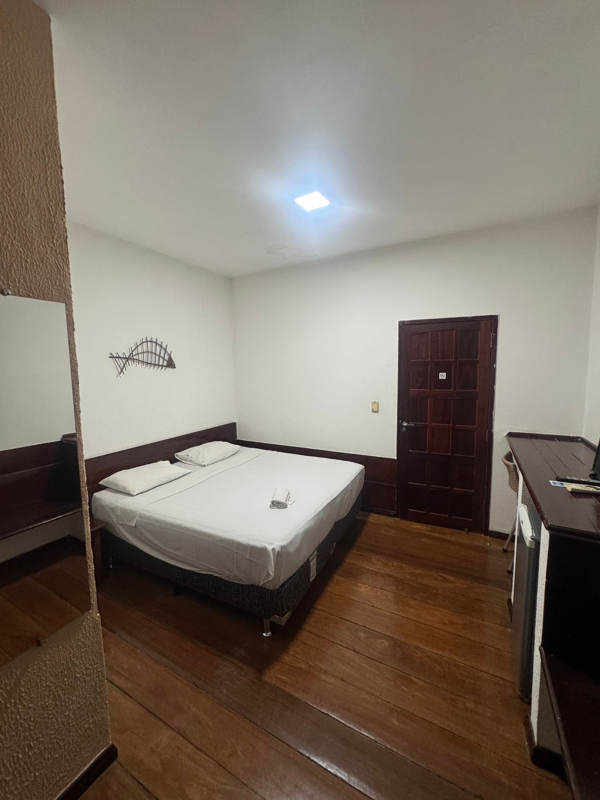 Quarto simples com duas camas, piso de madeira, paredes brancas e uma porta de madeira escura fechada.