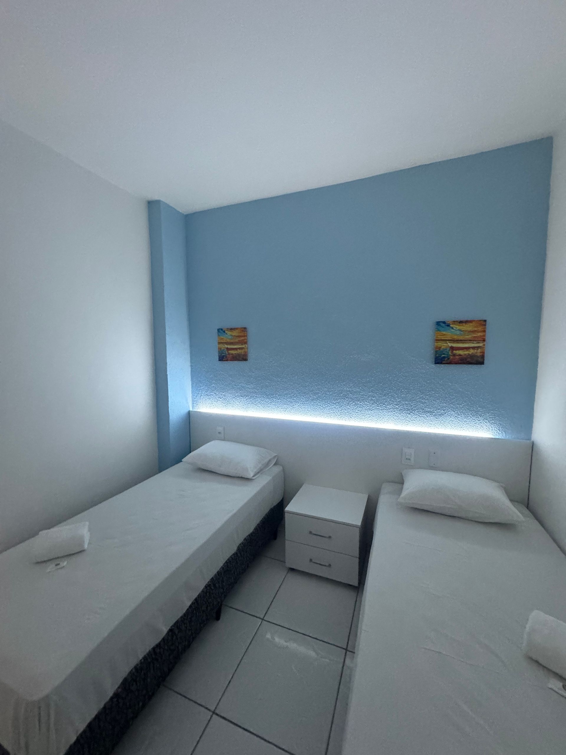 Quarto pequeno com duas camas de solteiro, paredes brancas, uma parede com detalhes em azul e uma faixa de luz suave acima da cabeceira.