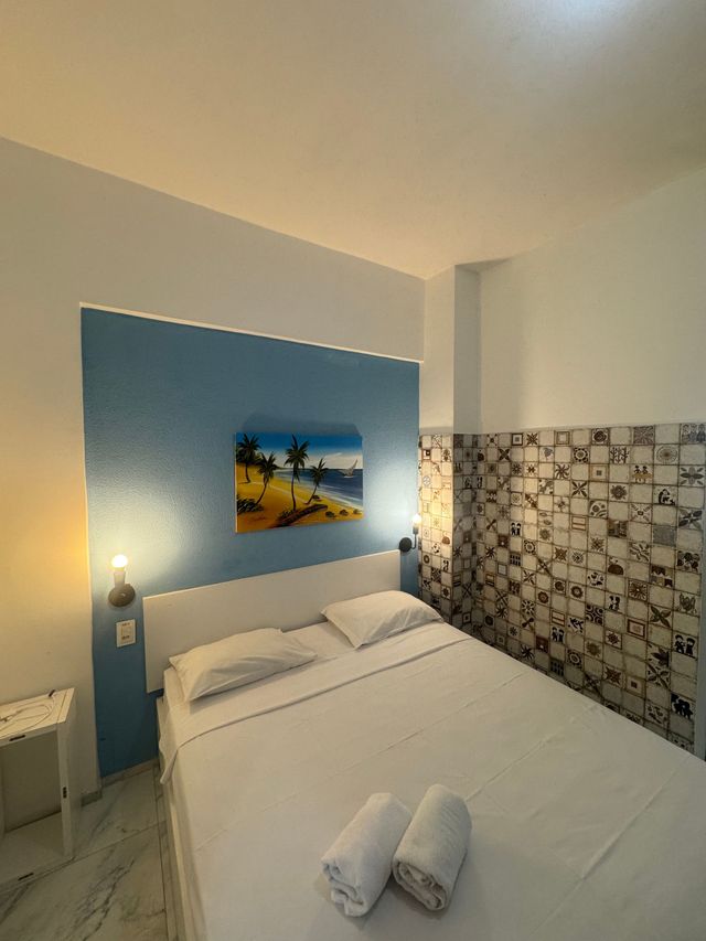 Cama de hotel impecavelmente arrumada com lençóis brancos, parede com detalhes em azul, obras de arte na parede e azulejos estampados em um quarto pequeno.