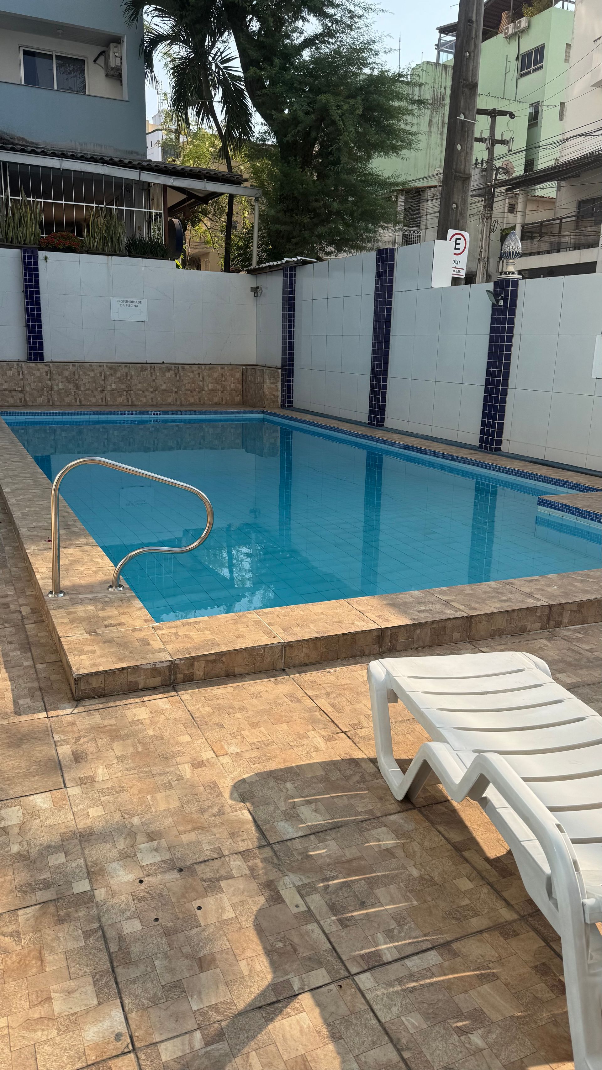 Pequena piscina externa com água azul, deck de azulejos, paredes brancas e uma espreguiçadeira em primeiro plano.