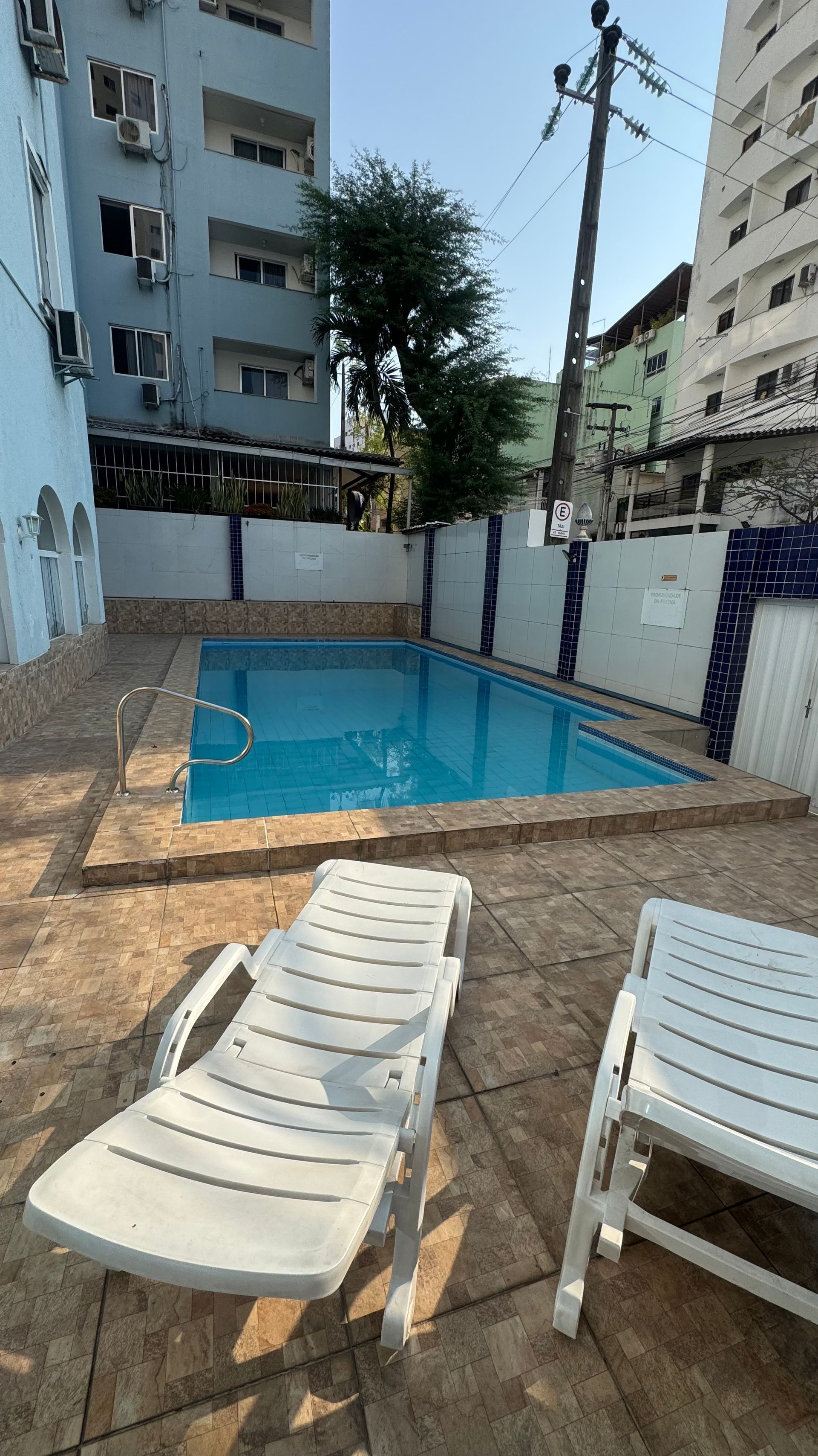 Pátio externo com uma pequena piscina azul, espreguiçadeiras brancas e prédios de apartamentos ao redor.