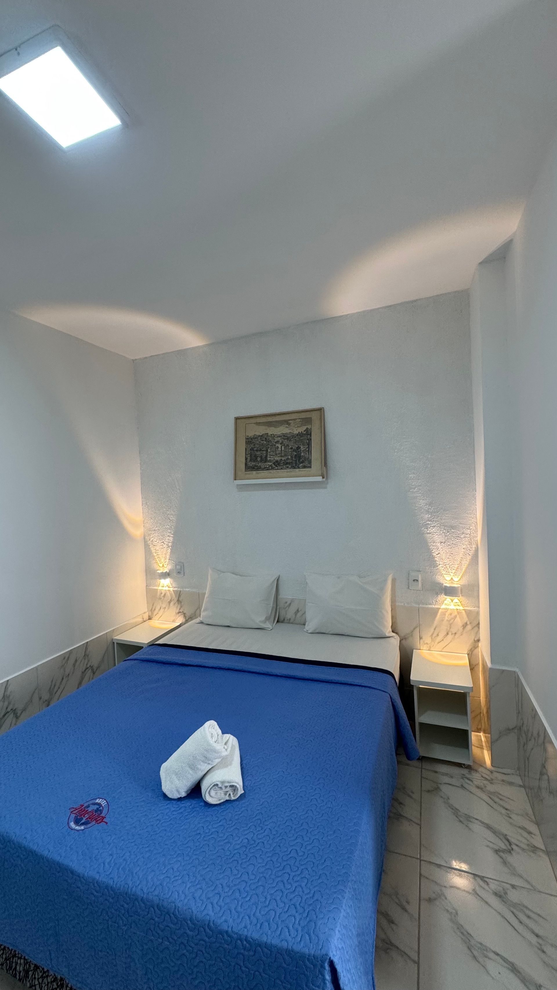 Quarto pequeno com colcha azul, dois abajures e um quadro na parede.