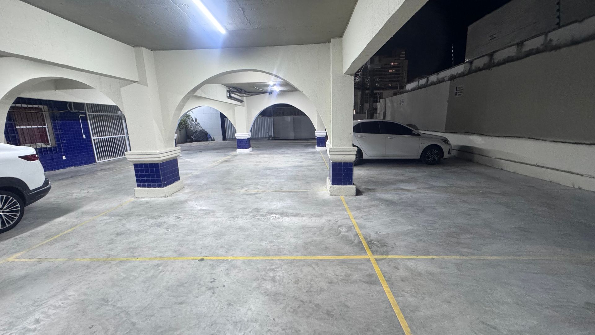 Garagem coberta com arcos de concreto, carros estacionados e marcações amarelas no piso.