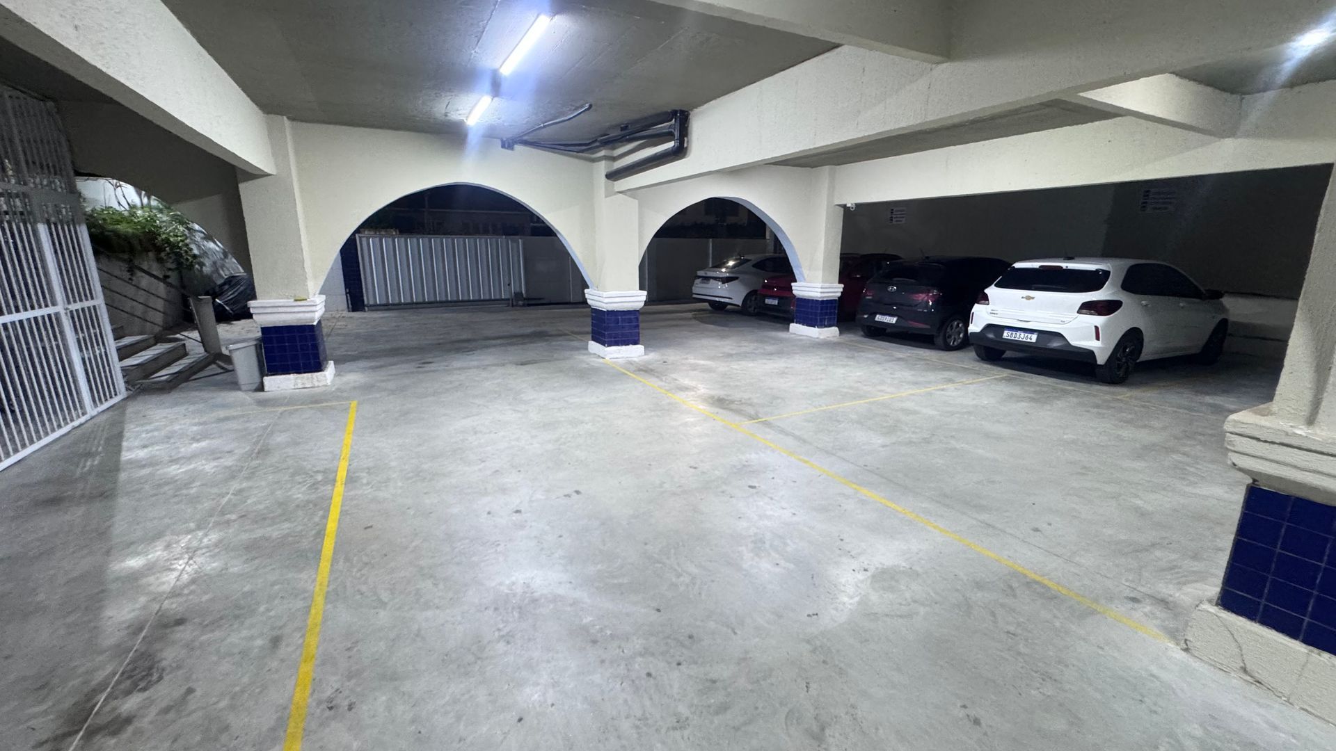 Garagem coberta com piso de concreto, aberturas em arco e vários carros estacionados sob luzes fortes.