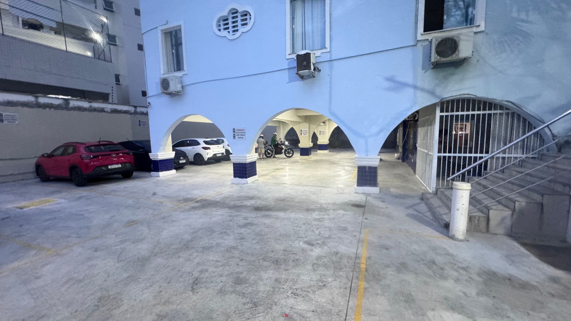 Pátio com prédio de apartamentos azul, aberturas em arco no térreo, um carro vermelho e escadas à direita.