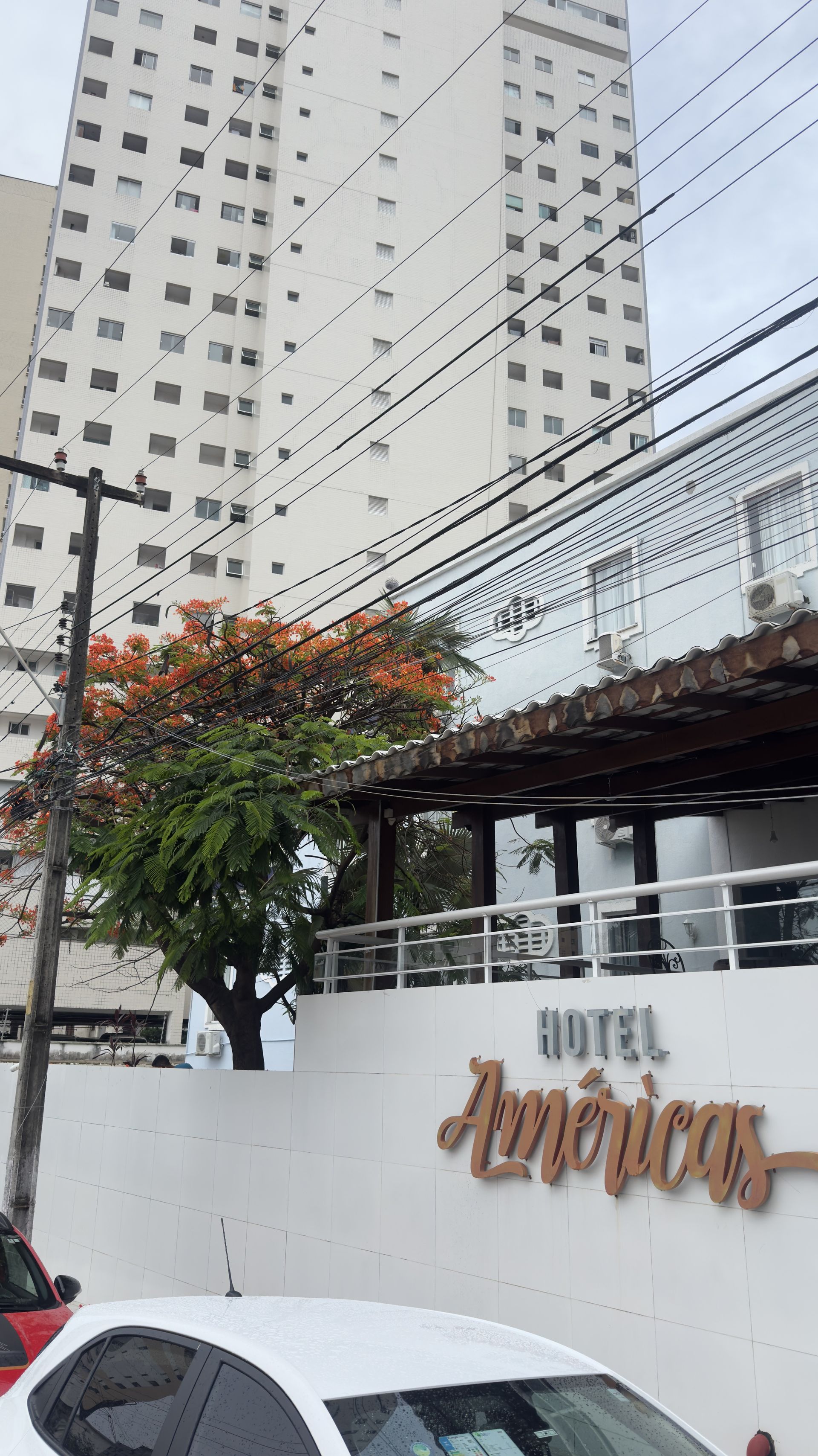 Placa do Hotel America em uma parede branca ao lado de uma rua, com prédios altos e uma árvore acima.