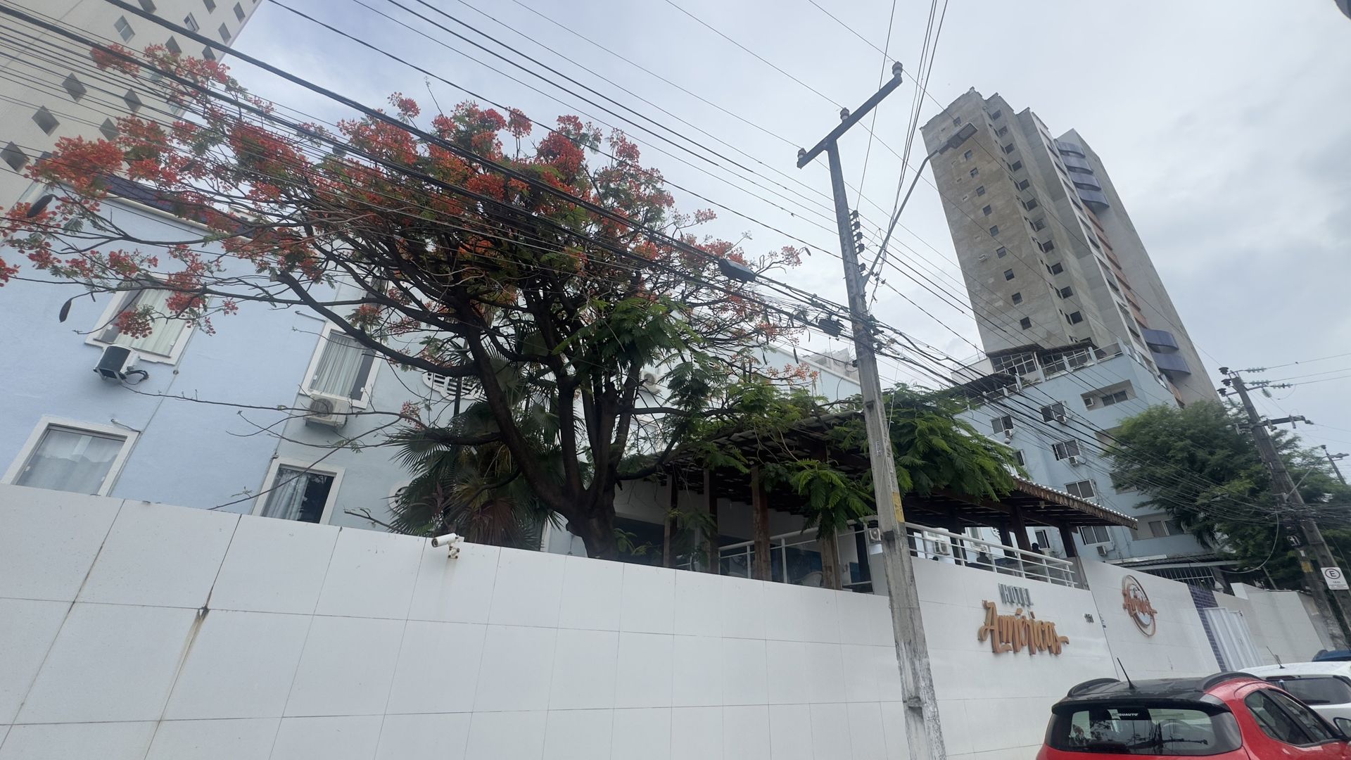 Parede branca e árvores na rua em frente a um prédio alto de apartamentos sob um céu nublado, com um carro vermelho na calçada.