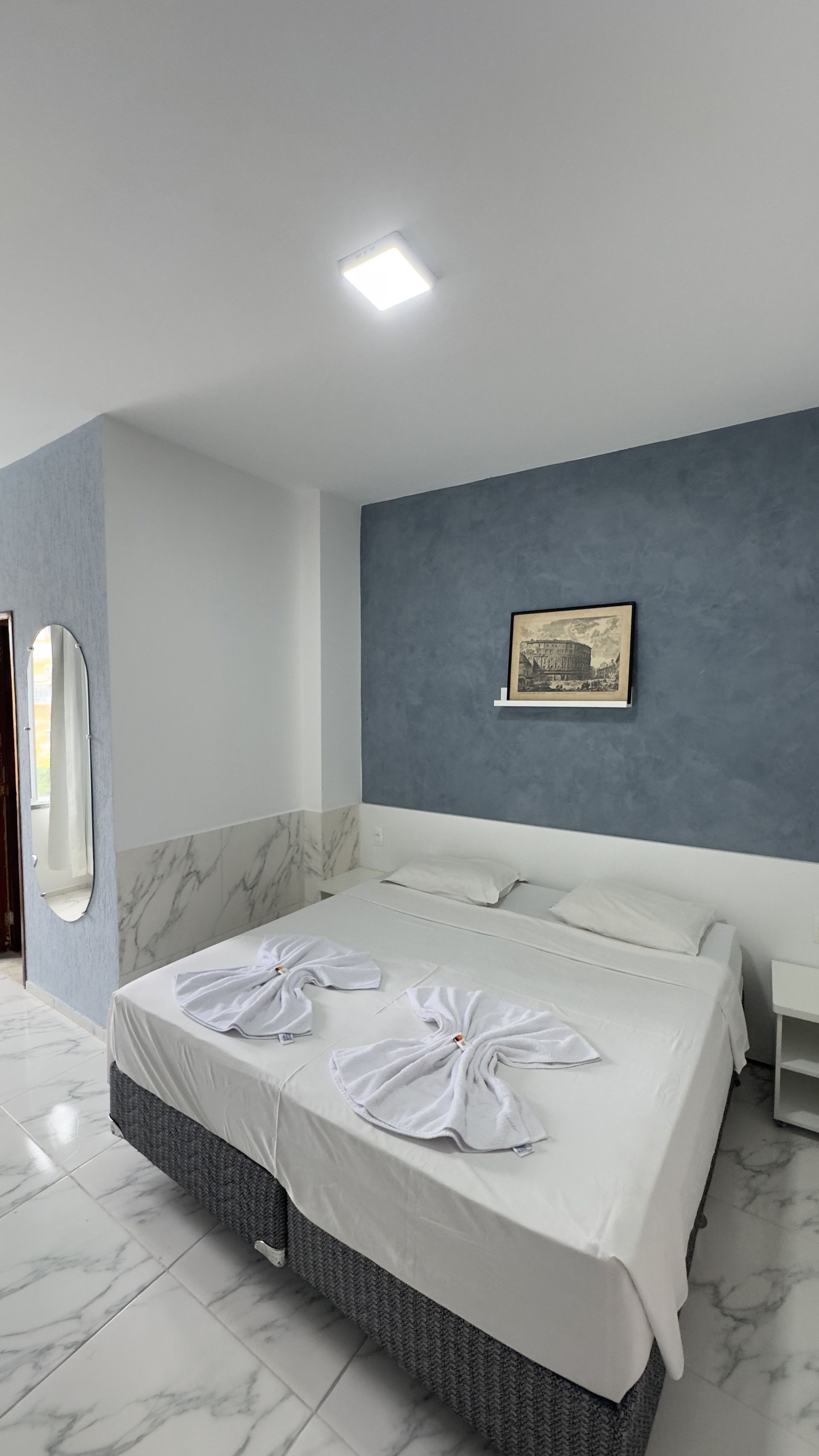 Quarto arrumado com cama branca, parede com detalhes em azul, obras de arte emolduradas e toalhas dobradas sobre a cama.